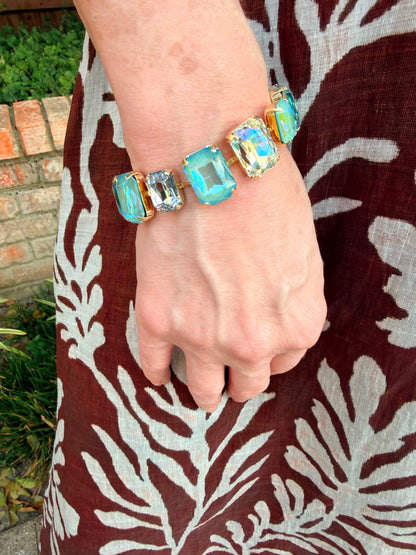 A close-up of a person's wrist wearing a chunky bracelet with five rectangular iridescent blue and gold cabochon stones, set against a brown and white patterned dress.