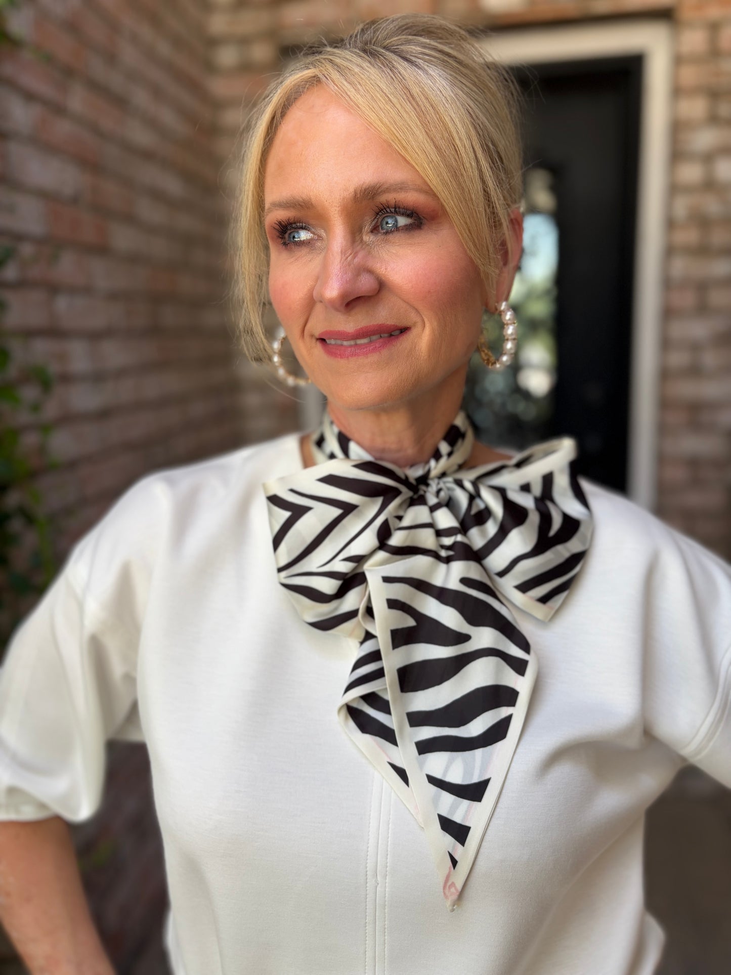 A woman wearing a white top and a black and white zebra-print neck scarf, standing in front of a brick wall.
