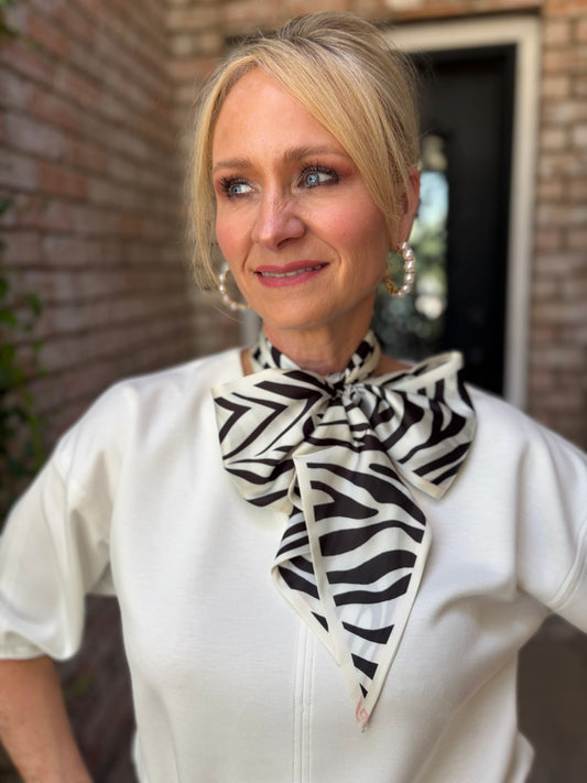 A woman wearing a white top and a black and white zebra-print neck scarf, standing in front of a brick wall.