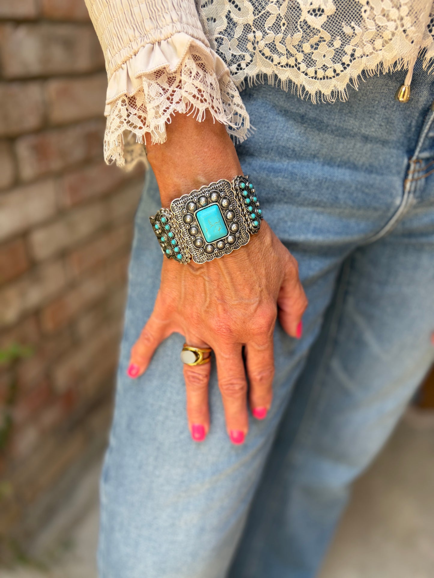 A close-up of a person's hand wearing a wide silver-toned cuff bracelet with a large turquoise stone and turquoise beads, paired with light blue jeans and a cream lace top.