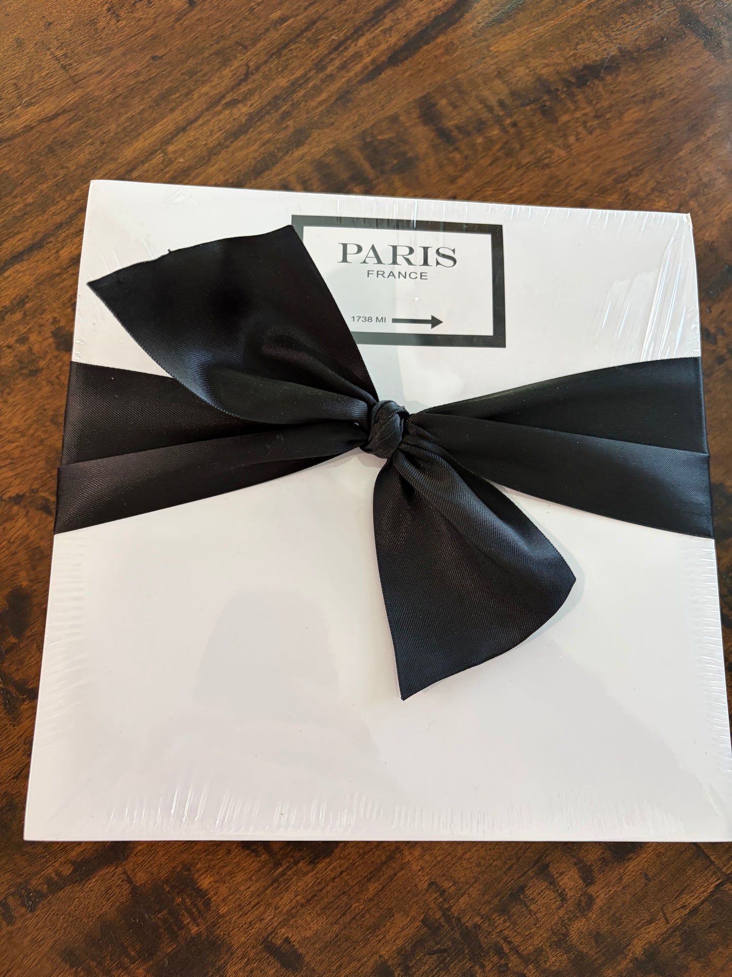 A white square notepad wrapped in clear plastic, tied with a large black satin bow, and featuring a label that reads 'PARIS FRANCE' on a dark wood surface.