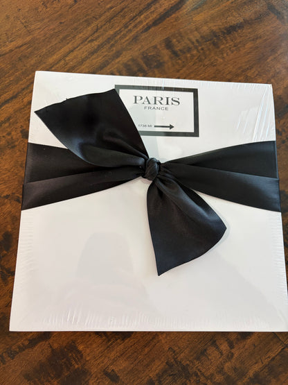 A white square notepad wrapped in clear plastic, tied with a large black satin bow, and featuring a label that reads 'PARIS FRANCE' on a dark wood surface.