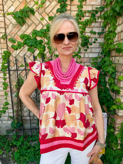 Woman wearing a colorful top with a floral pattern , red and pink and tan, flutter sleeve,  v neck aired with white denim