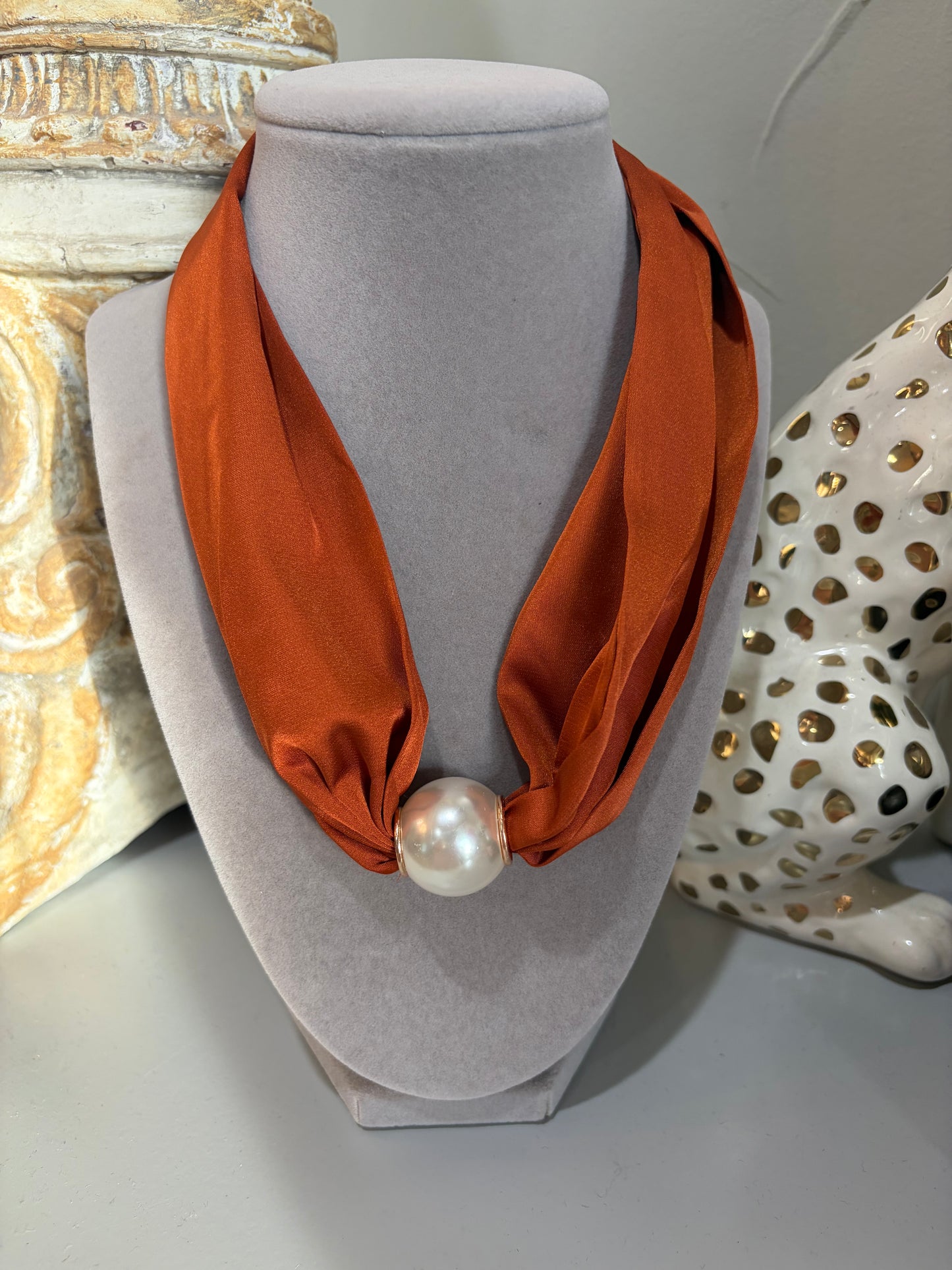 An orange silk scarf styled as a necklace on a gray velvet jewelry bust, featuring a large white pearl at the center, with decorative ceramic objects in the background.