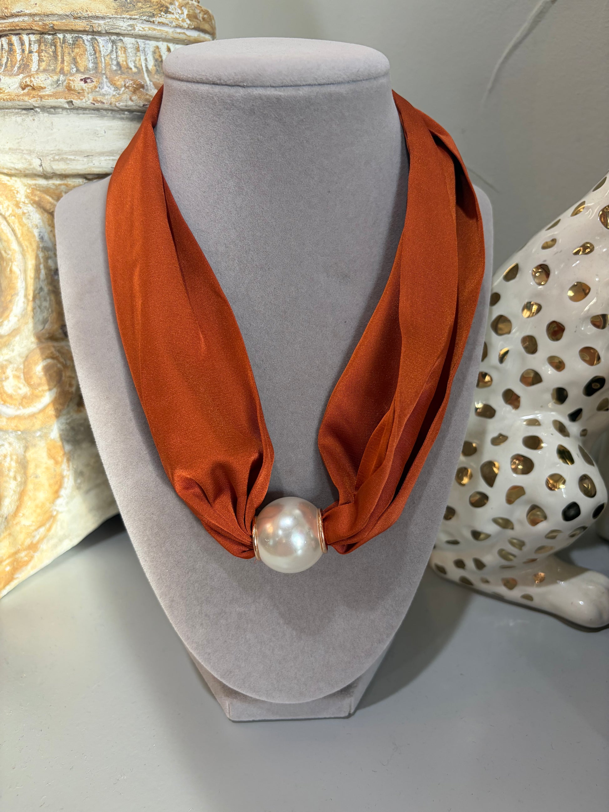 An orange silk scarf styled as a necklace on a gray velvet jewelry bust, featuring a large white pearl at the center, with decorative ceramic objects in the background.