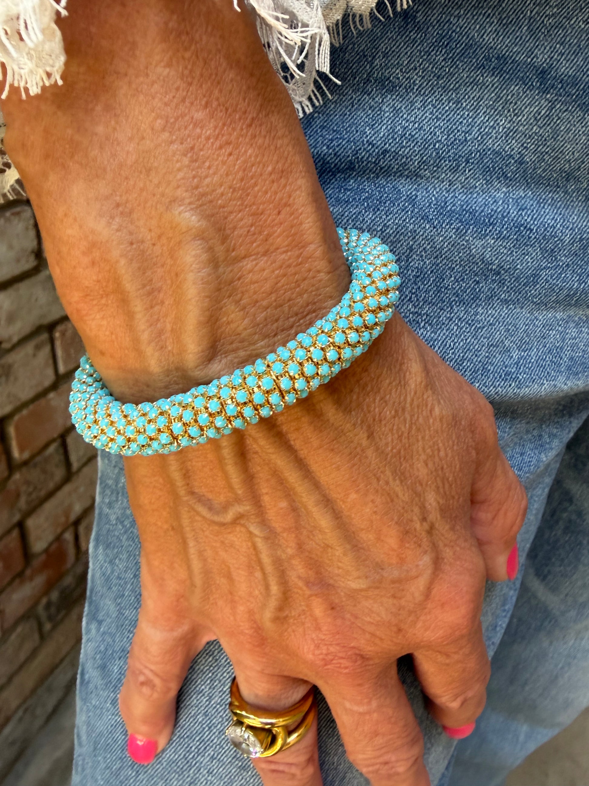 A close-up of a person's hand wearing a turquoise blue and gold beaded bracelet, resting on blue denim fabric.