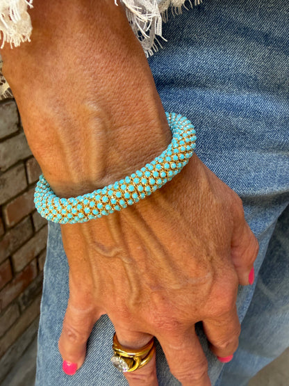 A close-up of a person's hand wearing a turquoise blue and gold beaded bracelet, resting on blue denim fabric.