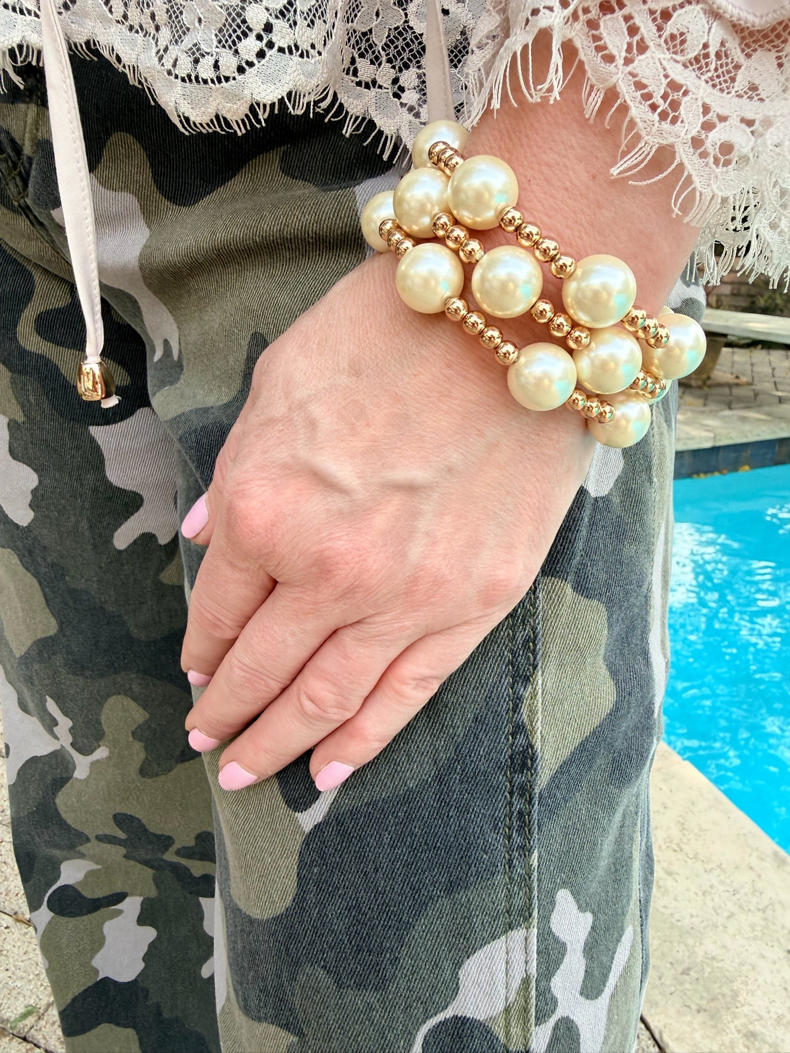 A close-up of a person's hand and wrist wearing a three-strand beaded bracelet with large cream pearl-like beads and gold-toned spacer beads, paired with camouflage pants and a lace-trim top near a pool.