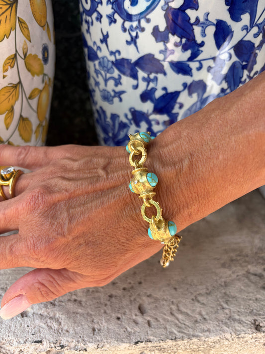 A close-up of a person's hand and wrist wearing a gold chain bracelet with turquoise-colored oval accents, set against a patterned blue-and-white garment and a textured gray wall.