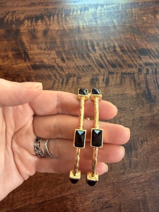 A pair of gold-toned drop earrings with rectangular black gemstone accents, held in a person's hand against a dark wood background.