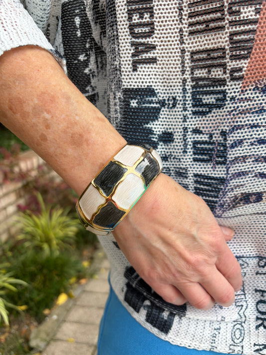 A close-up of a person's wrist wearing a chunky geometric cuff bracelet with alternating white and black square panels outlined in gold.