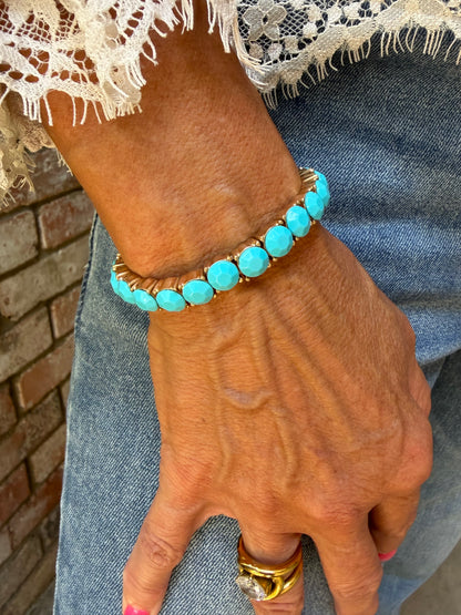 A turquoise and gold beaded bracelet worn on a person's wrist, shown with a hand resting on blue jeans.