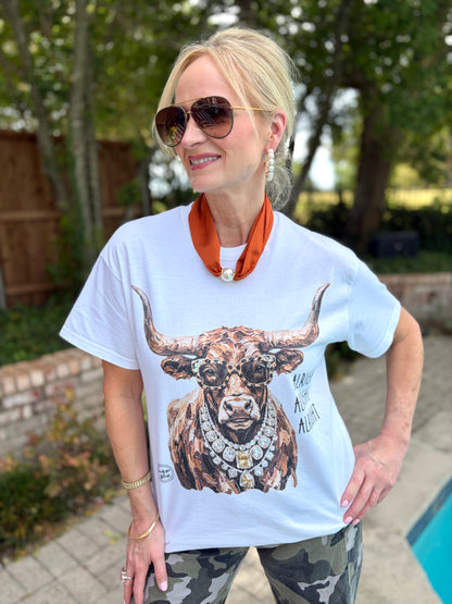 A woman wearing a white graphic t-shirt with a detailed longhorn  illustration and an orange neck scarf, standing outdoors near a pool.