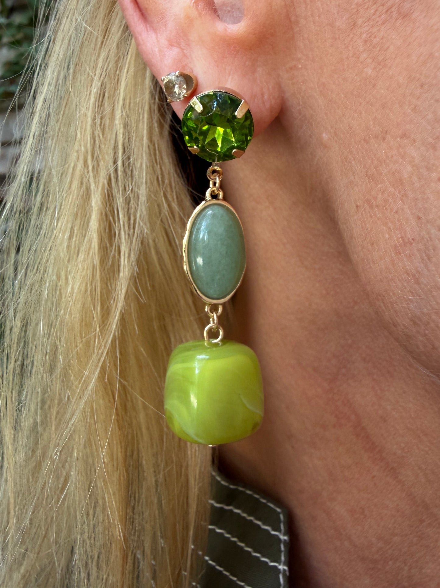 A close-up of a person's ear wearing a drop earring with a faceted green top stone, a light green oval stone, and a larger lime green rounded drop, all in gold-tone settings. water resistant