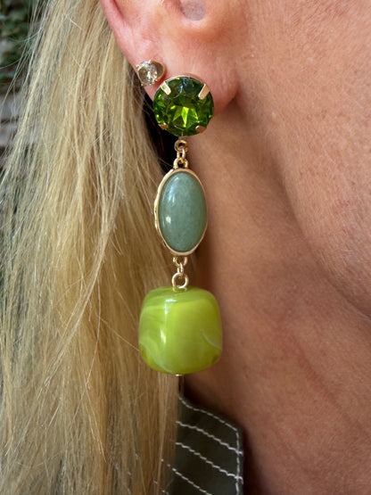 A close-up of a person's ear wearing a drop earring with a faceted green top stone, a light green oval stone, and a larger lime green rounded drop, all in gold-tone settings. water resistant