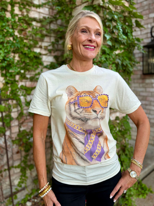 A smiling blonde woman wearing a cream-colored graphic t-shirt with a large print of a tiger wearing purple sunglasses and a purple bandana. She is posing outdoors in front of a brick wall with green ivy.