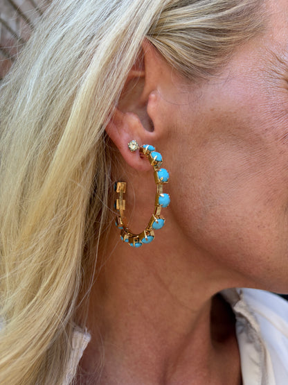 A close-up of a woman's ear wearing a gold hoop earring adorned with small turquoise blue beads and a small clear stud earring.