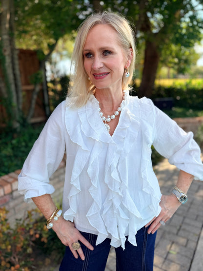 A woman wearing a white long-sleeve blouse with multiple layers of ruffles, paired with dark blue jeans and jewelry, standing outdoors in a garden setting.