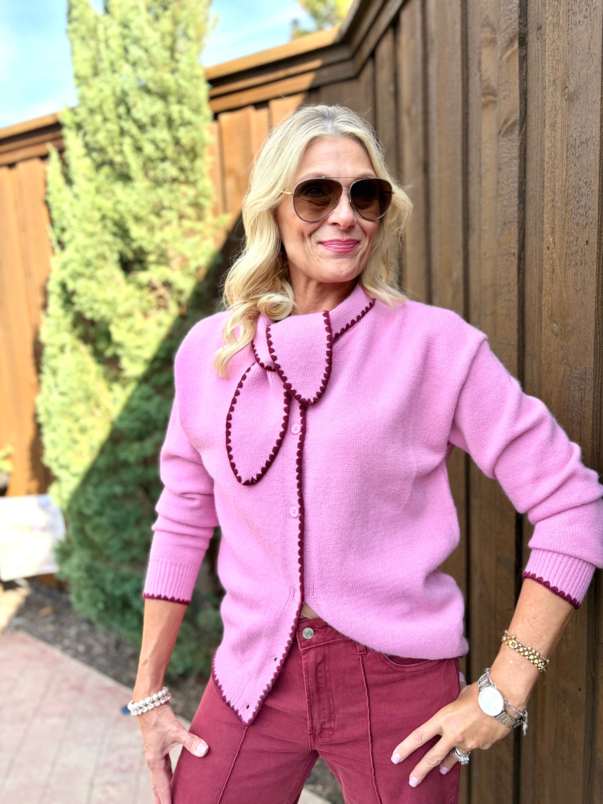 A woman wearing a light pink knit cardigan button down  with dark berry trim with tie around neck line and matching pink pants, standing outdoors in front of a wooden fence.