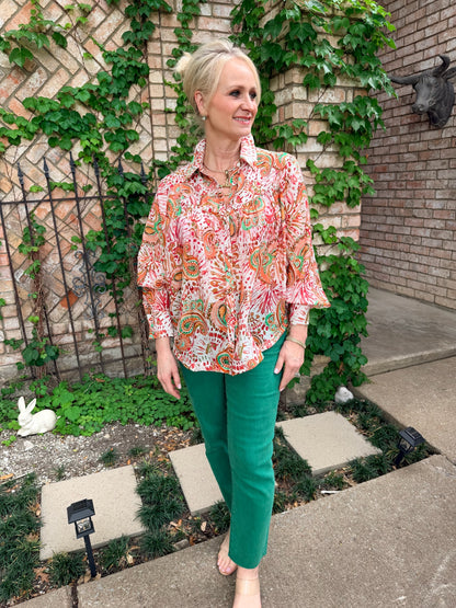 Woman wearing a patterned long sleeve blouse with oranges, greens, button down, and green denim pants 