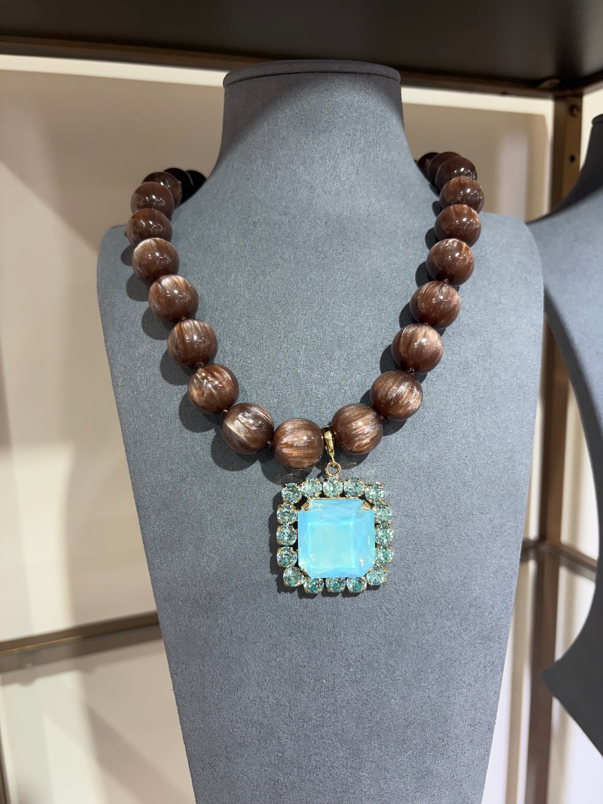 A chunky statement necklace displayed on a gray mannequin bust. The necklace features a strand of polished dark brown beads with a central square pendant.