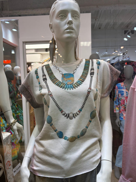 A mannequin wearing a cream-colored t-shirt with a colorful graphic print of three layered necklaces, set inside a clothing store.