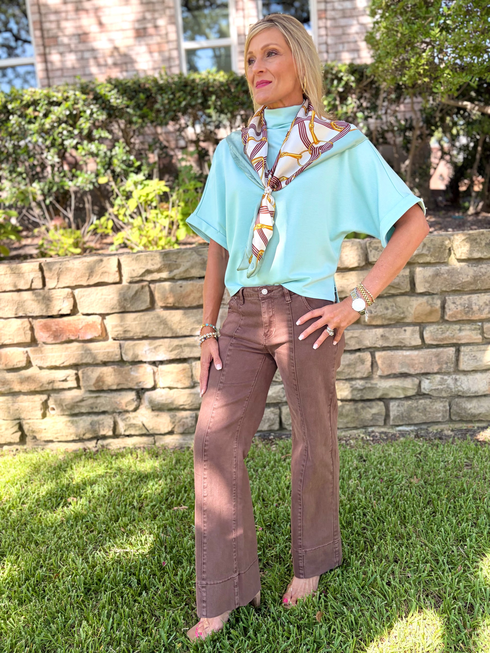 A woman stands outdoors on a grassy lawn in front of a stone wall, wearing a light aqua short-sleeve blouse with a multicolor patterned scarf tied around the neck, brown wide-leg pants