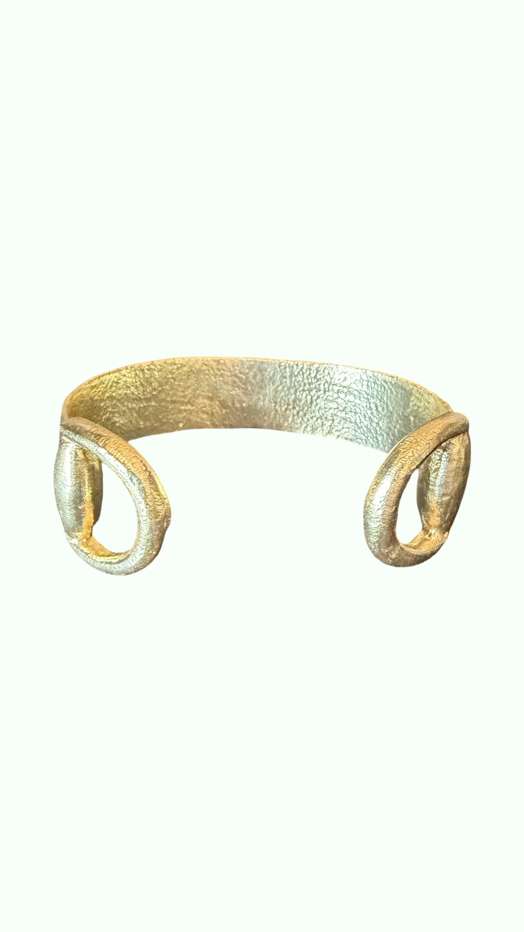 A textured gold cuff bracelet with two oval buckle like details on the ends, shown against a plain off-white background.