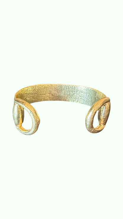 A textured gold cuff bracelet with two oval buckle like details on the ends, shown against a plain off-white background.