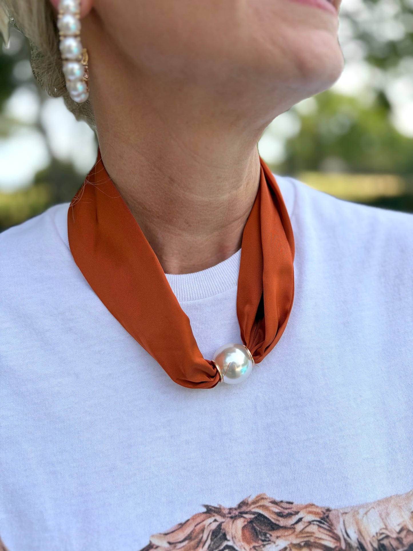A close-up lifestyle shot of a woman wearing a rust-orange satin neck scarf tied in a knot with a single white pearl, styled over a white t-shirt.