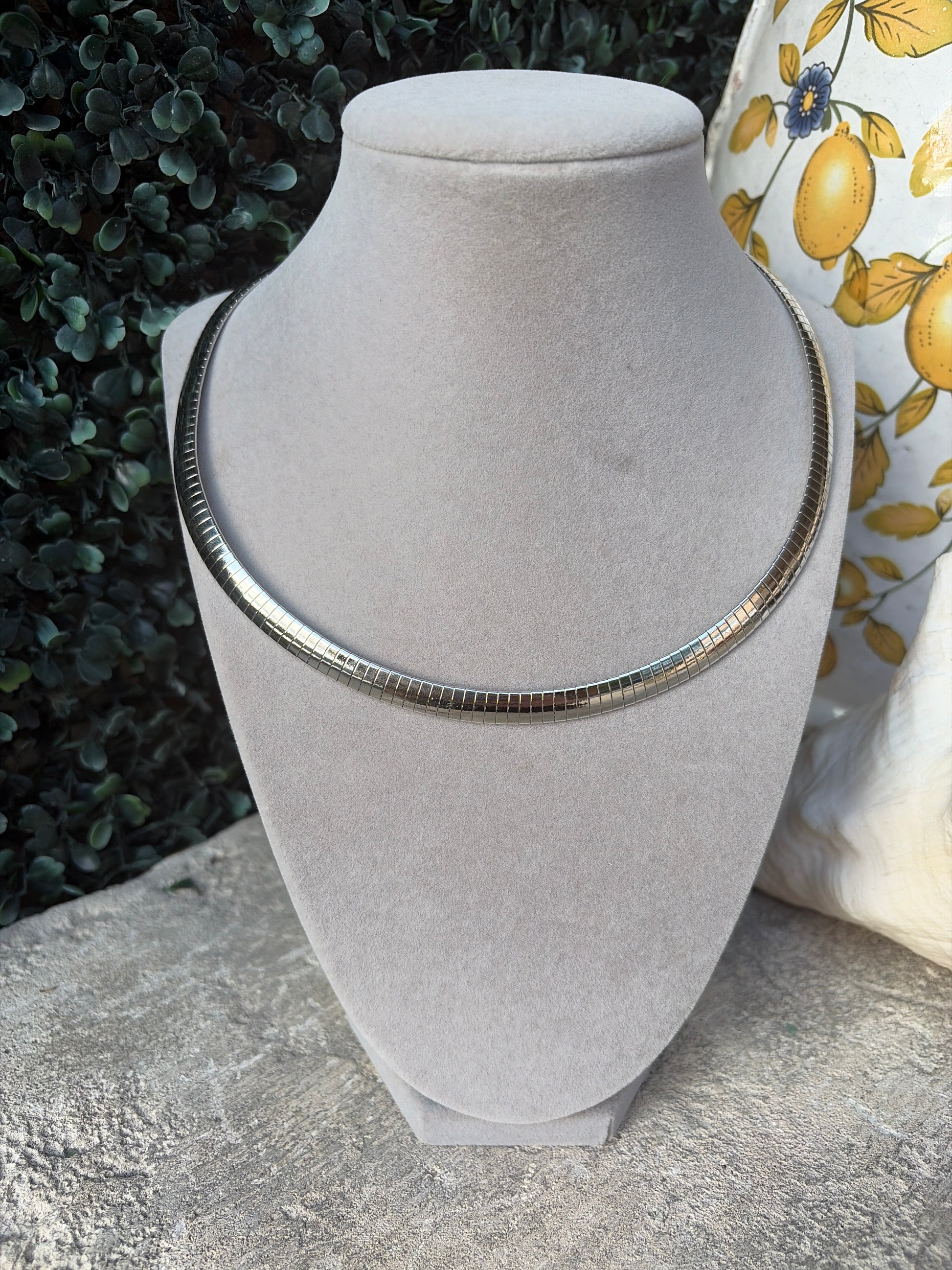 A silver-toned snake chain necklace displayed on a light gray velvet jewelry bust, set against a background of green foliage and a yellow lemon-patterned fabric.
