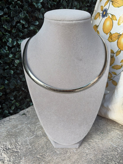 A silver-toned snake chain necklace displayed on a light gray velvet jewelry bust, set against a background of green foliage and a yellow lemon-patterned fabric.