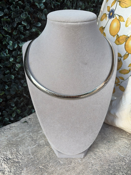 A silver-toned snake chain necklace displayed on a light gray velvet jewelry bust, set against a background of green foliage and a yellow lemon-patterned fabric.