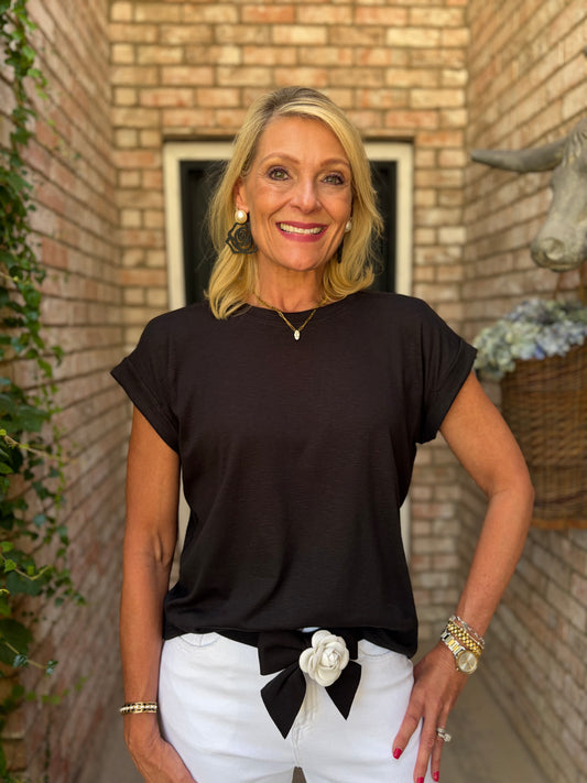 A smiling blonde woman wearing a black short-sleeve  simple tee and white pants with a black bow and white flower detail, standing in front of a brick wall.