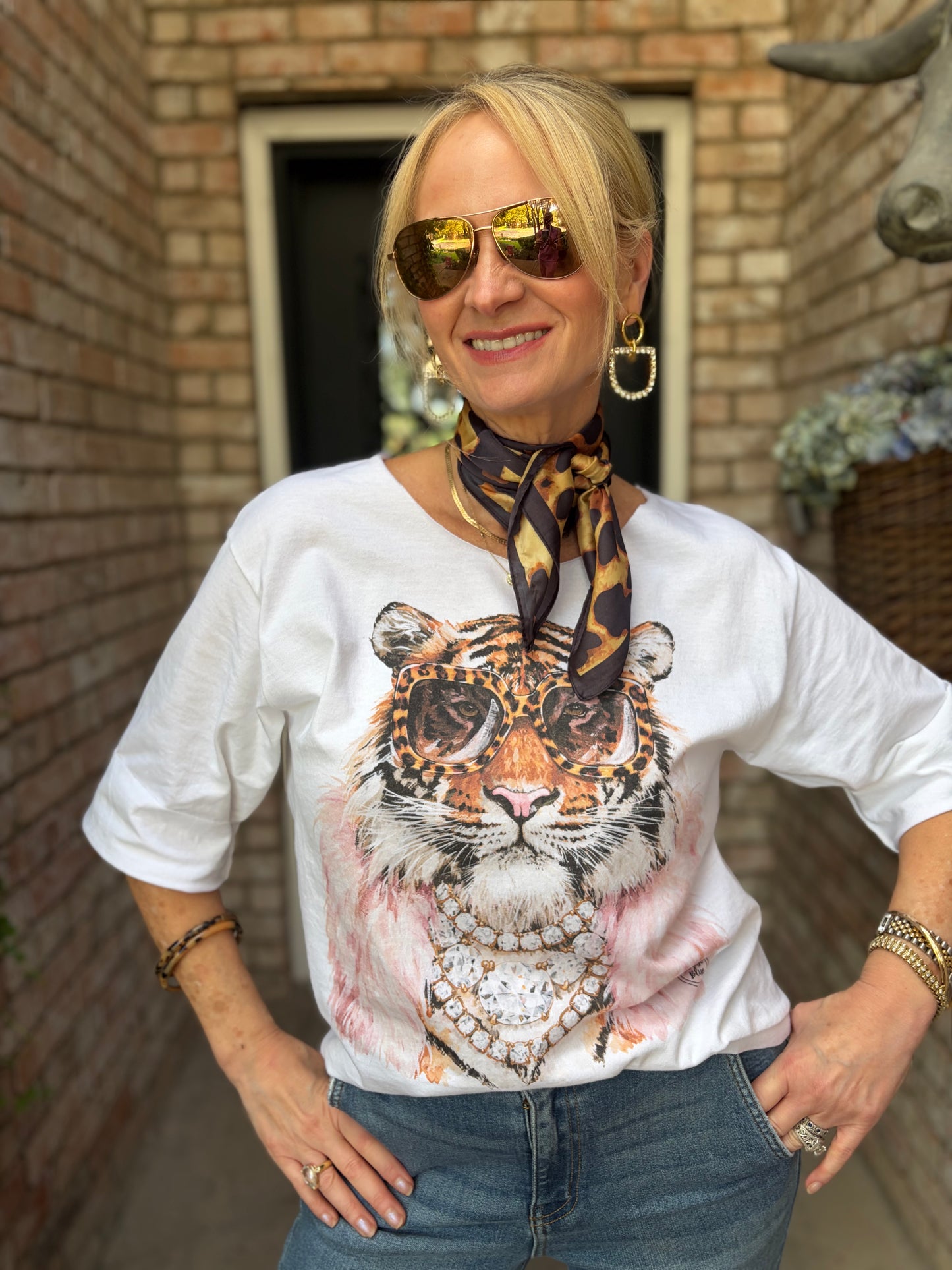 A woman wearing a white graphic t-shirt with a large tiger face print, styled with a leopard-print neck scarf, sunglasses, and jewelry, standing in front of a brick wall.