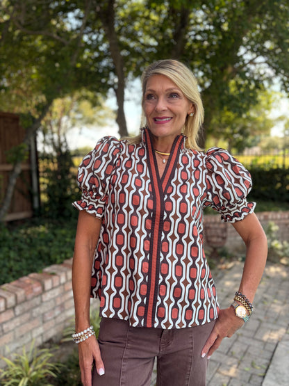 A short, puff sleeved Entro top that is v neck with faux collar in geometric design of rust, crwam and brown detail 
