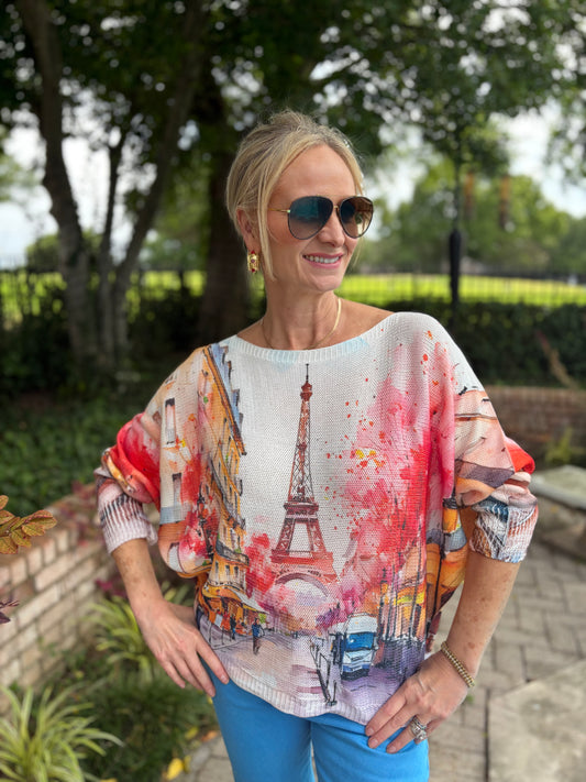 A woman wearing a colorful Paris-themed printed top with the Eiffel Tower and street scene, paired with blue pants, standing outdoors in a garden setting.