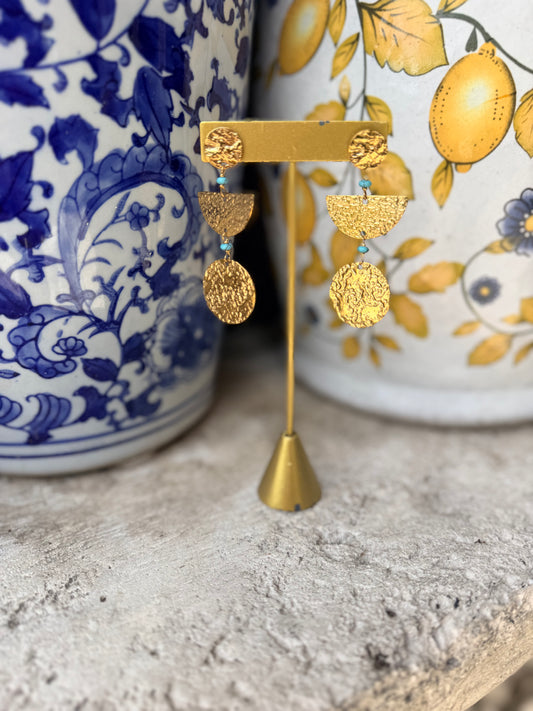 Gold statement earrings displayed on a gold stand, featuring textured circular studs and layered semi-circle and oval drops with small turquoise blue bead accents, set against a rustic stone surface with blue and yellow patterned ceramic vases in the background.