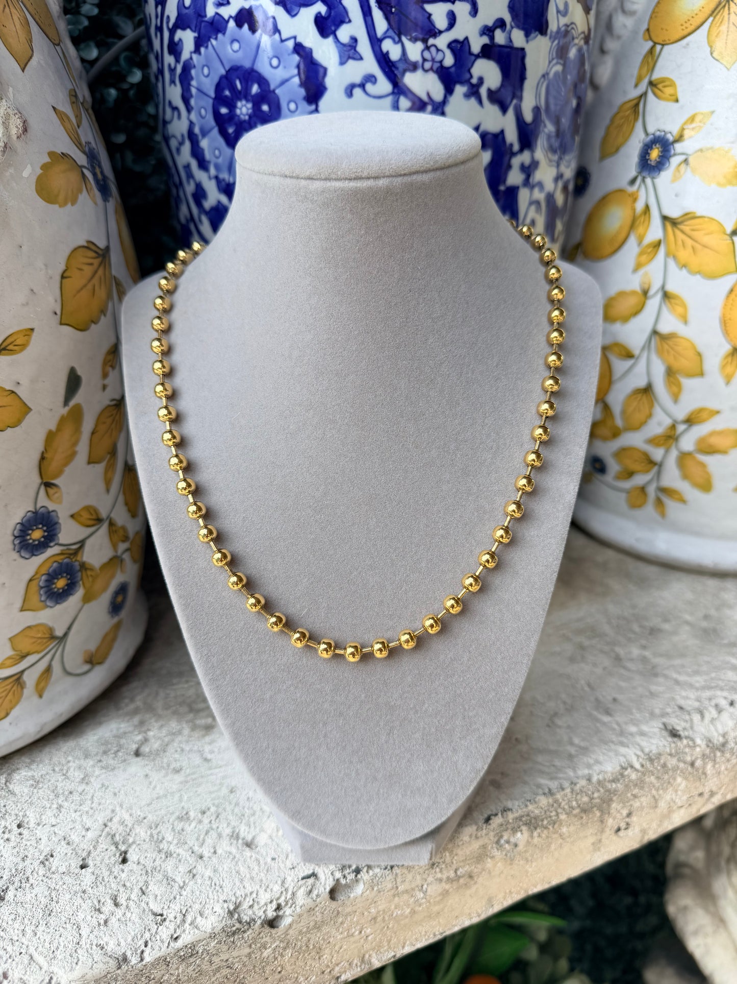 A delicate gold beaded necklace displayed on a light gray velvet jewelry bust, set against a rustic background with blue and yellow patterned ceramic vases.
