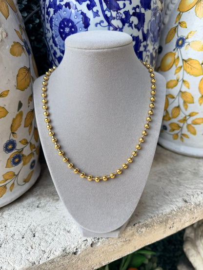 A delicate gold beaded necklace displayed on a light gray velvet jewelry bust, set against a rustic background with blue and yellow patterned ceramic vases.
