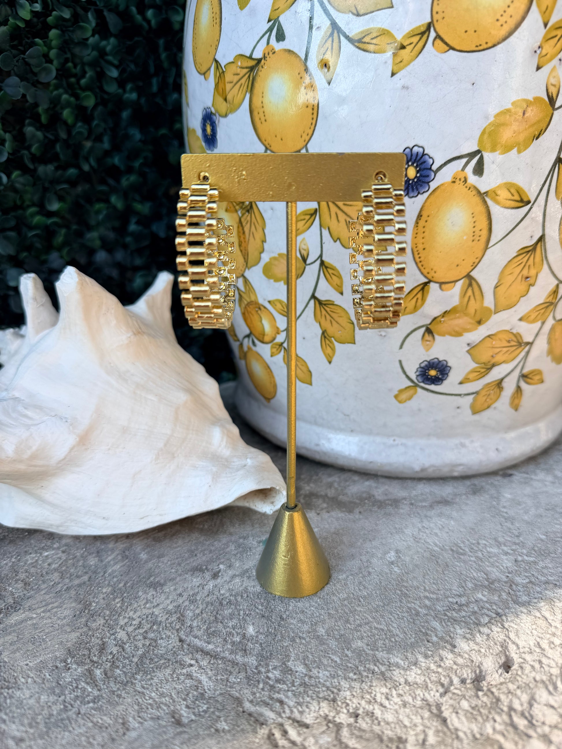 A pair of chunky gold-tone chain-link hoop earrings displayed on a small gold stand, set against a decorative white ceramic vase with a yellow lemon print and a white seashell on a stone surface.