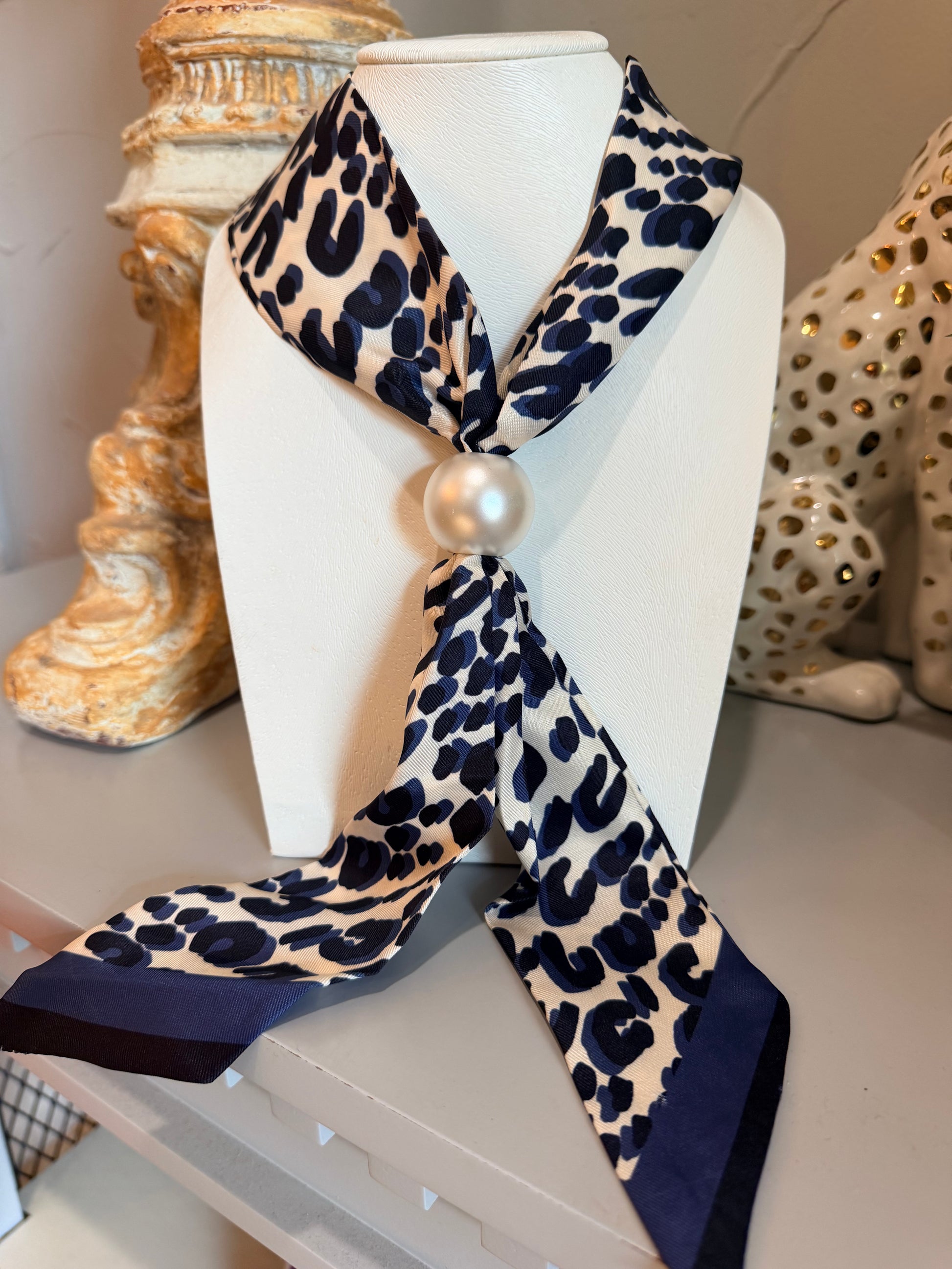 A leopard print scarf with navy blue spots and a solid navy border, styled on a white mannequin bust with a faux pearl at the knot, set against a background of decorative ceramic figurines.