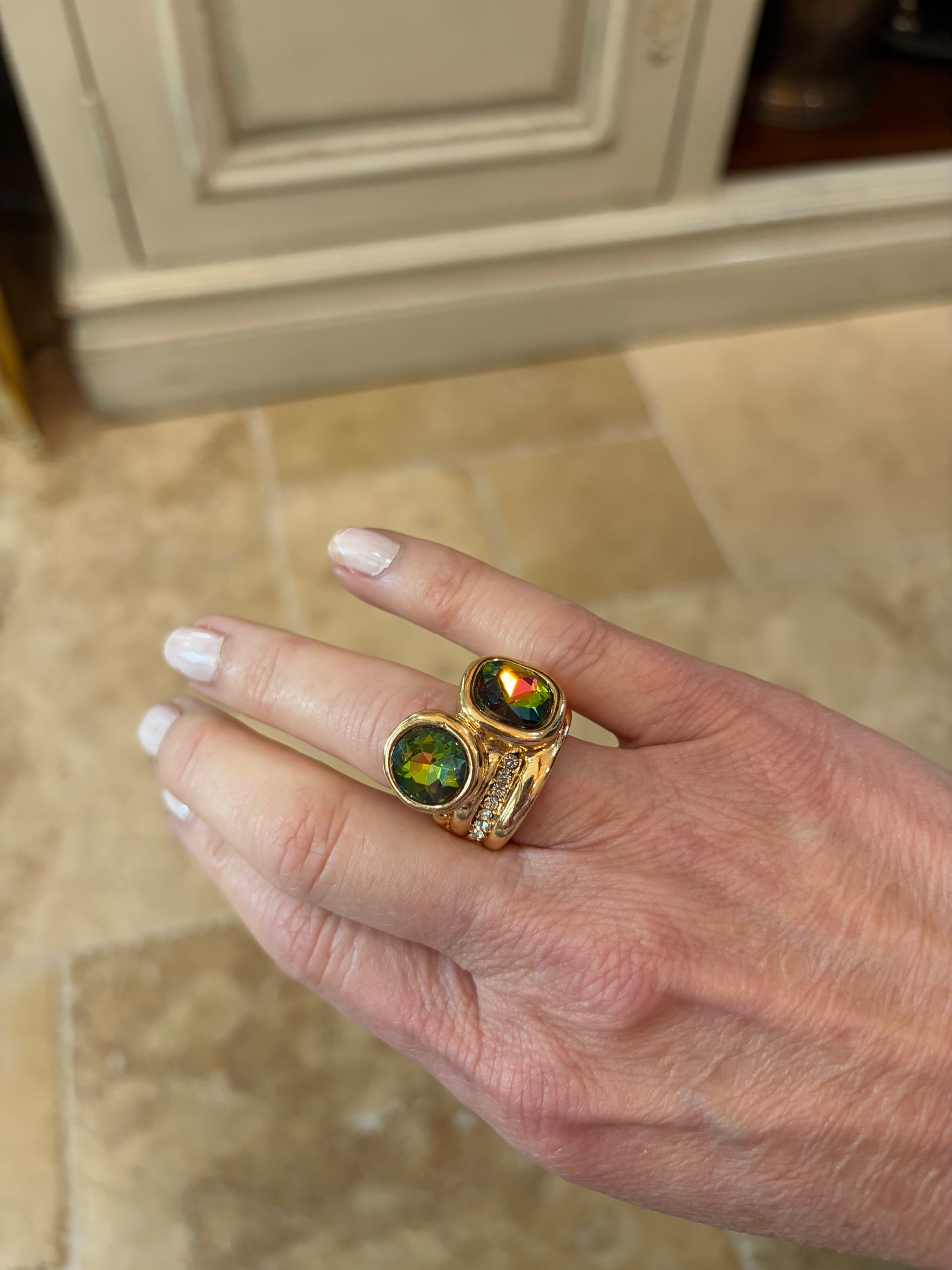 Hand wearing a gold ring with a colorful gemstone on a tiled floor.