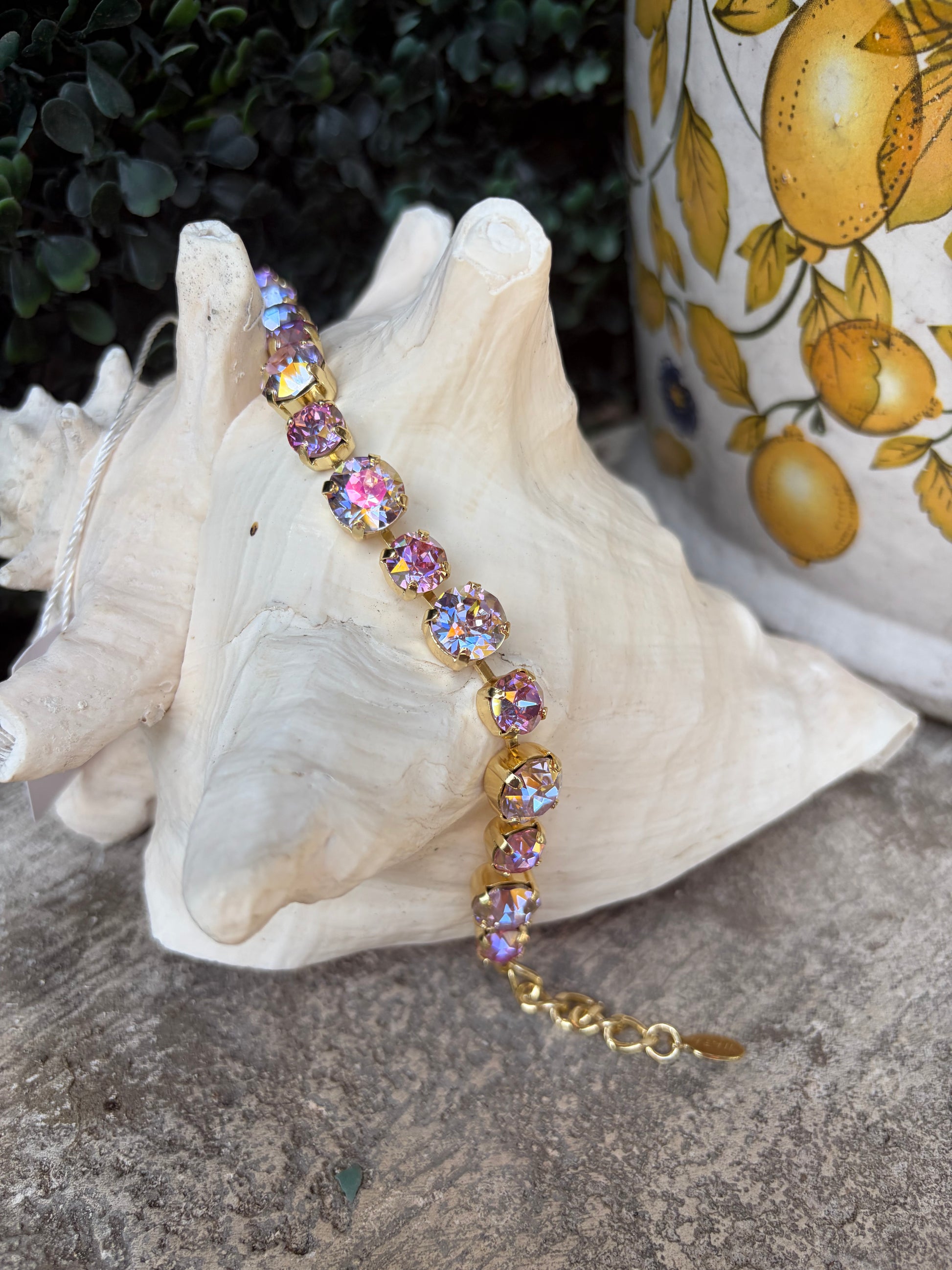 A gold-tone bracelet with iridescent pink and white rhinestones draped over a large white seashell, with a lemon-patterned ceramic pot in the background.