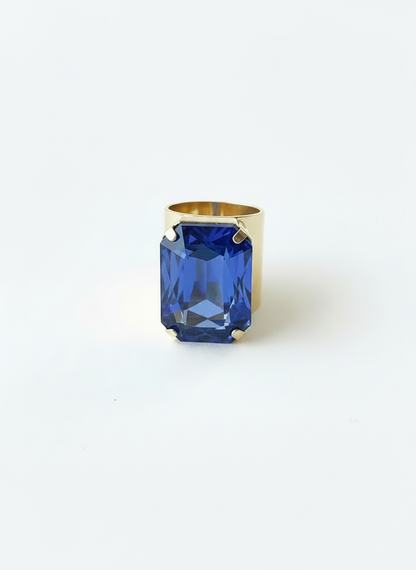 A gold ring featuring a large, faceted blue rectangular gemstone, photographed against a seamless white background.