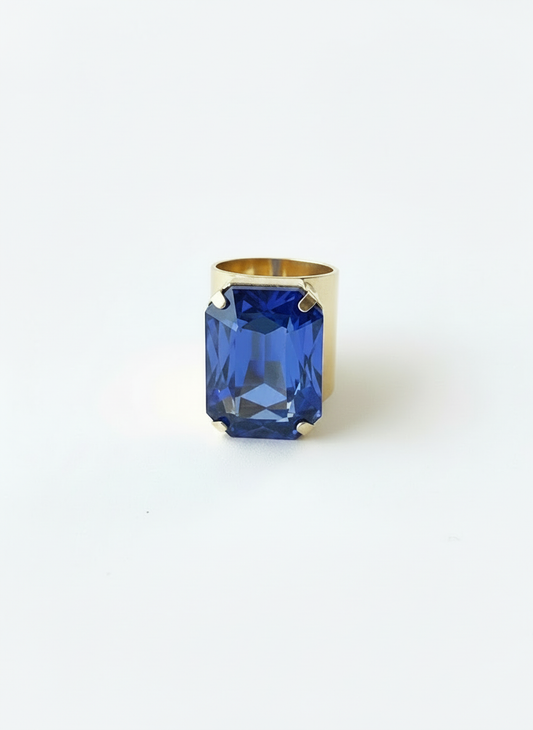 A gold ring featuring a large, faceted blue rectangular gemstone, photographed against a seamless white background.