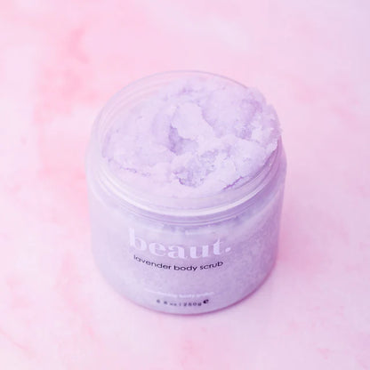 A clear plastic jar of beaut. lavender body scrub filled with light purple, crystalline scrub, set against a soft pink background.
