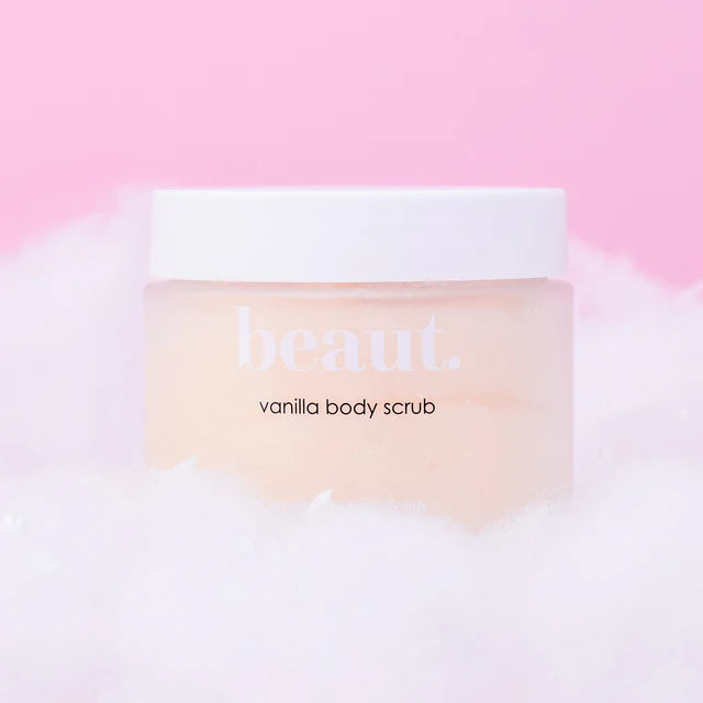 A jar of beaut. vanilla body scrub with a white lid and pale peach-colored scrub visible inside, resting on white foam against a soft pink background.