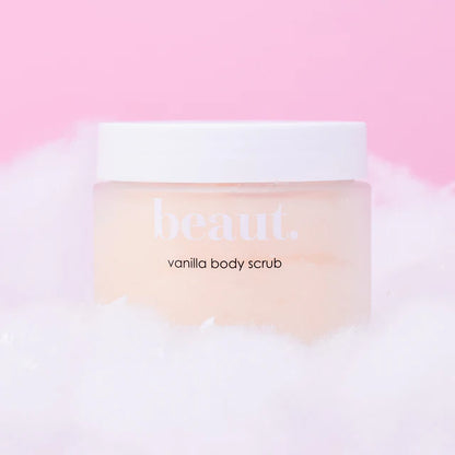 A jar of beaut. vanilla body scrub with a white lid and pale peach-colored scrub visible inside, resting on white foam against a soft pink background.