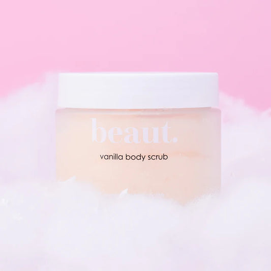 A jar of beaut. vanilla body scrub with a white lid and pale peach-colored scrub visible inside, resting on white foam against a soft pink background.
